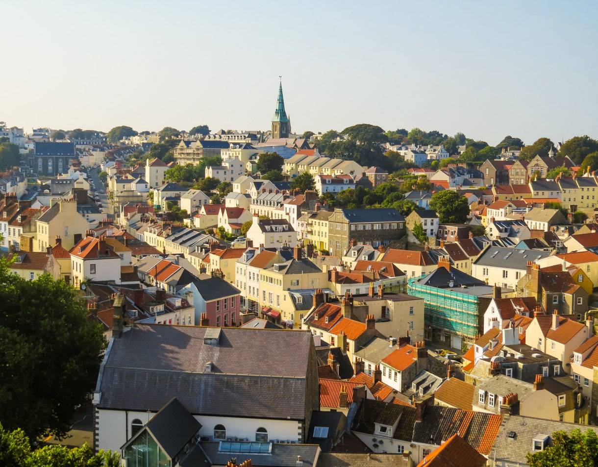 An elevated view shows a dense array of historic buildings with red-tiled roofs stretching across the hillside town of St. Peter Port under a clear sky.