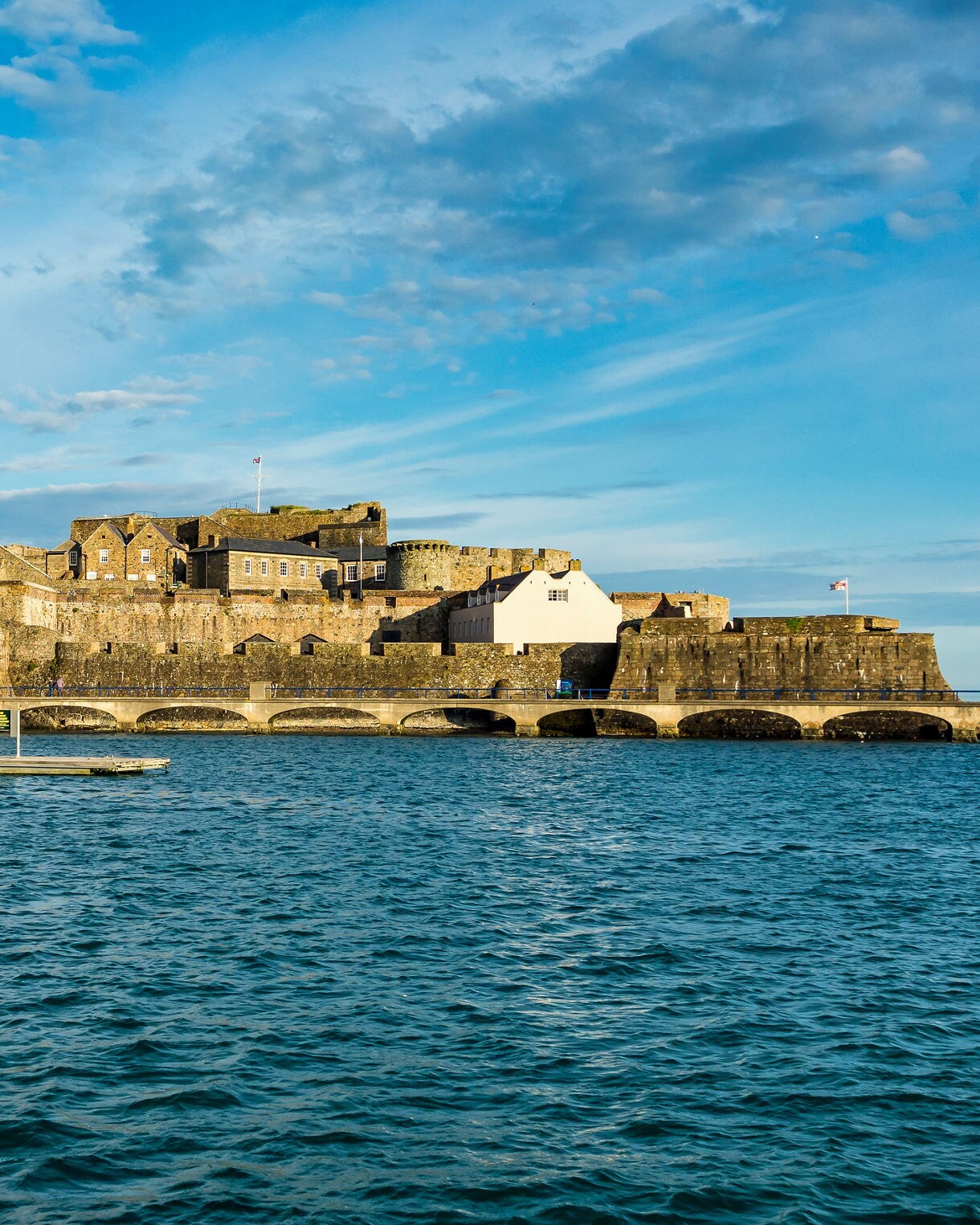A historic stone fortress complex built on an island structure sits offshore in deep blue waters beneath a sky filled with light clouds.