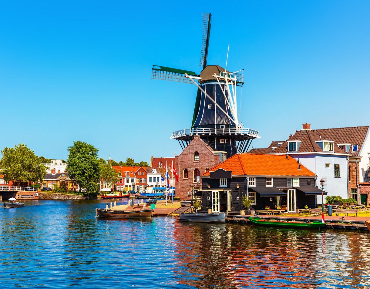 Historic windmill standing beside a canal in Haarlem, surrounded by traditional Dutch houses with red roofs, moored boats and trees reflected in the water under a bright blue sky.