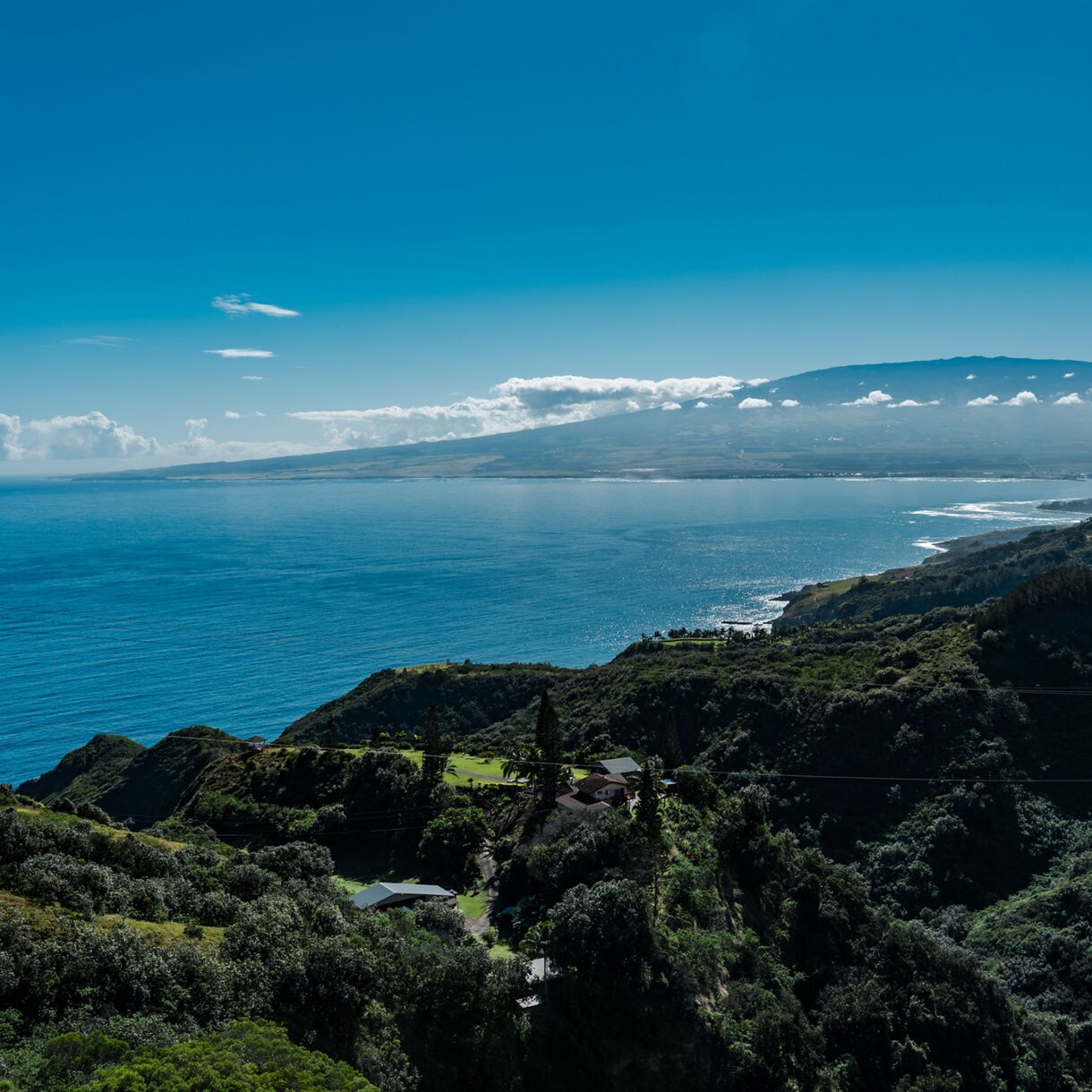 Panoramic view from Haleakalā’s slopes overlooking the Pacific Ocean, with rolling green hills and a distant mountain under a clear blue sky.