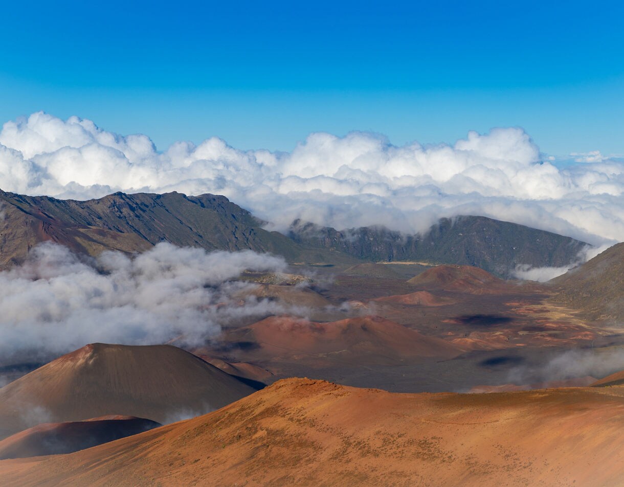 Panoramic view of Haleakalā Crater with red volcanic slopes, misty clouds and distant mountain ridges under a clear blue sky.