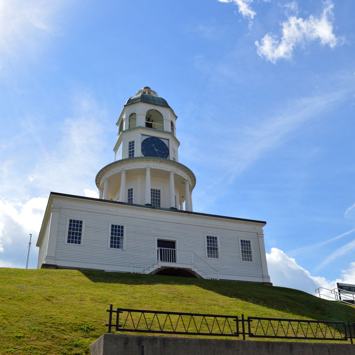 Low-angle view of the Halifax Town Clock atop a grassy hill, its white exterior and domed top set against a vivid blue sky with scattered clouds.