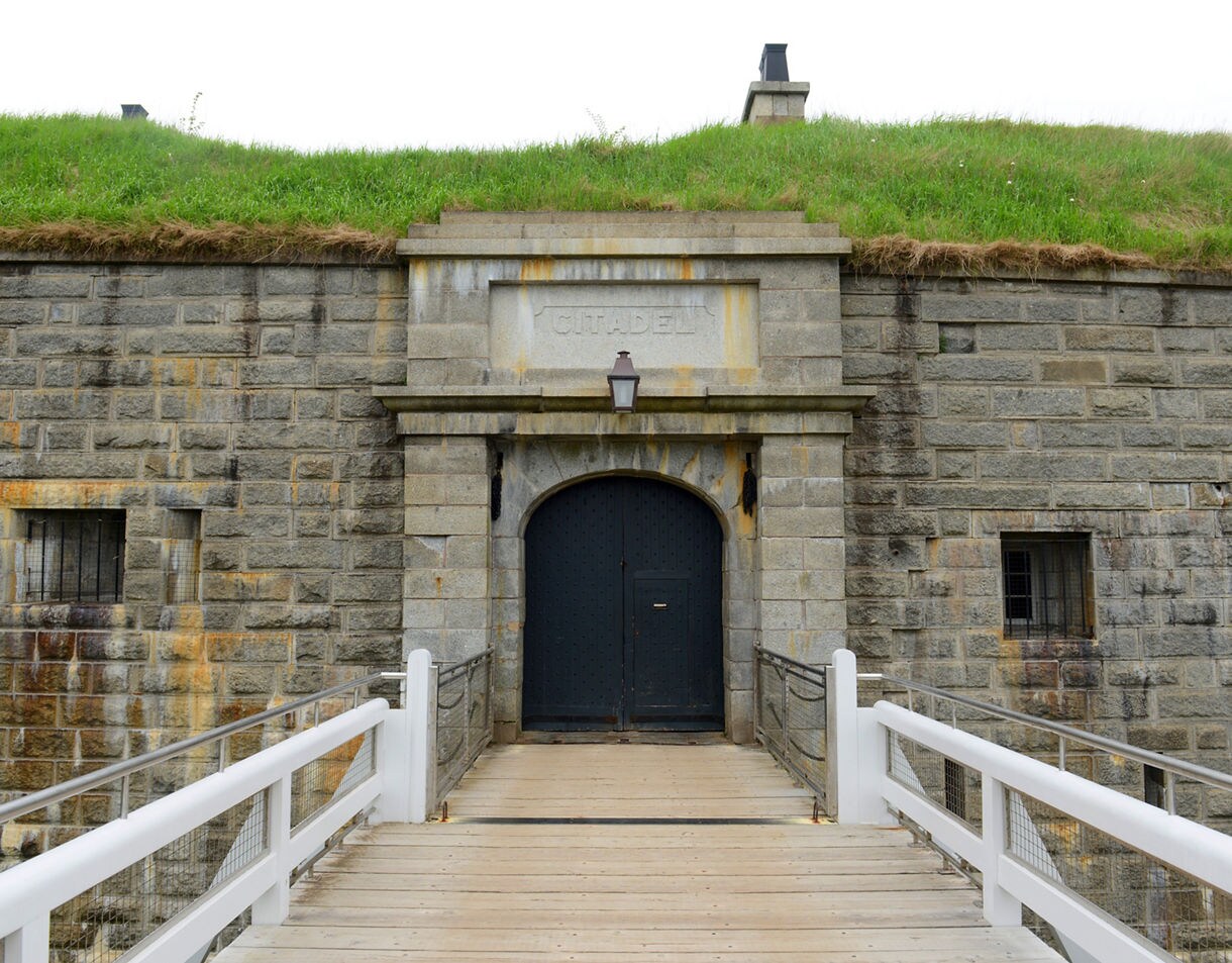 Stone entrance of Halifax Citadel with a wooden footbridge leading to a large black door set into thick grey walls topped with grassy earthworks.