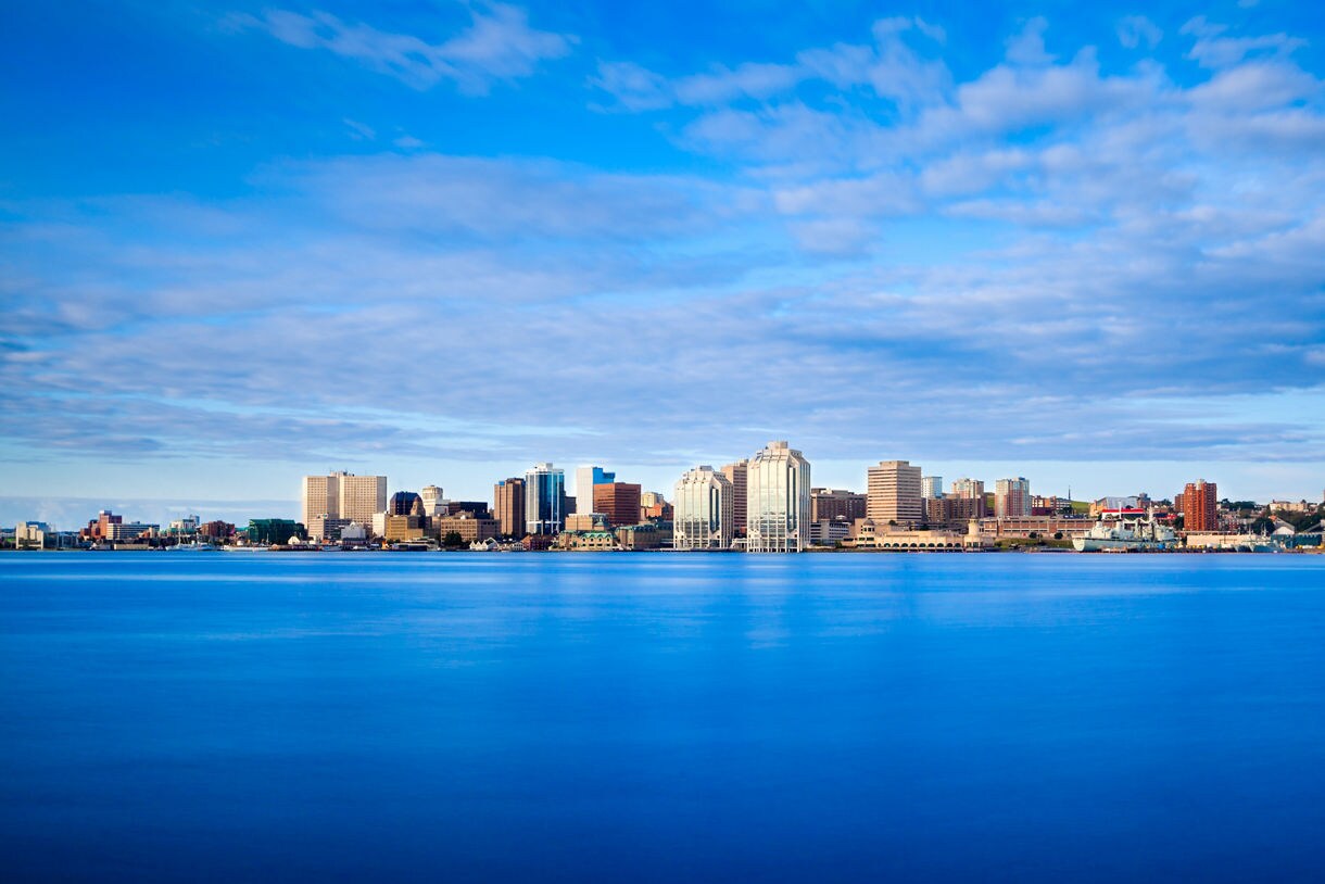 Wide view of Halifax’s waterfront skyline with modern high-rises reflecting over a smooth blue harbor under a partly cloudy sky.