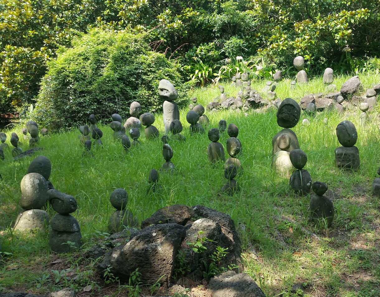 tone statues shaped from volcanic rock scattered across a grassy clearing at Hallim Park on Jeju Island, surrounded by lush green foliage.