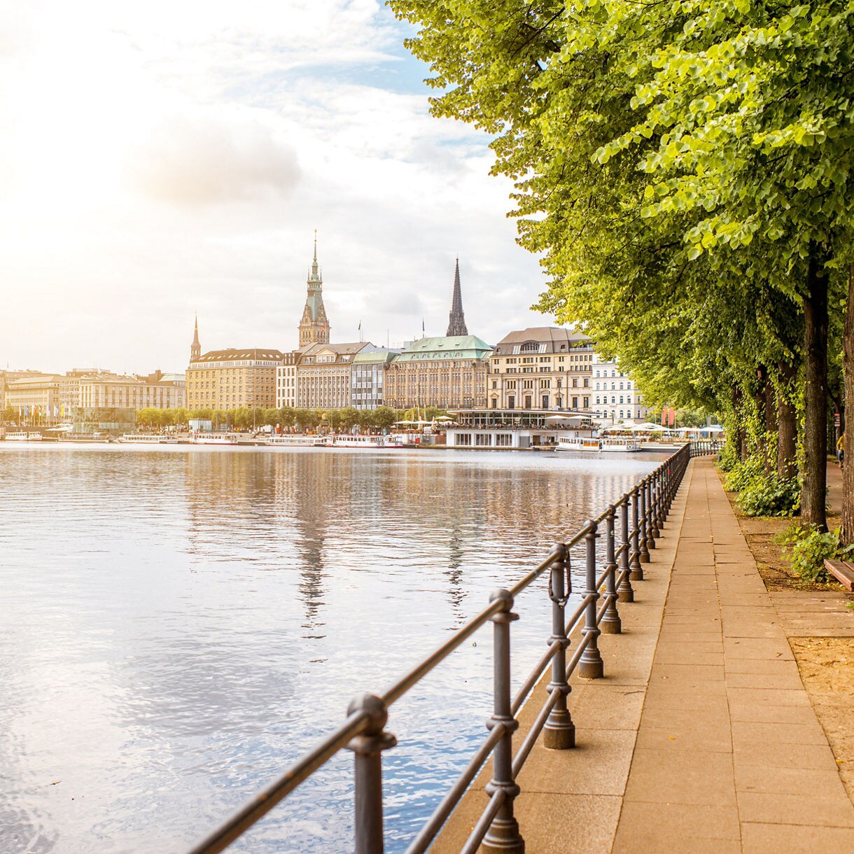 Scenic view of Hamburg’s Inner Alster Lake with calm water, leafy promenade and city skyline featuring church spires in the background.