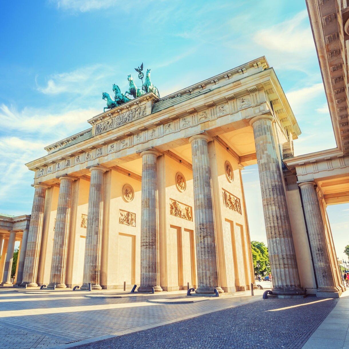 The Brandenburg Gate in Berlin with tall neoclassical columns, detailed carvings and a bronze quadriga statue under a bright blue sky.