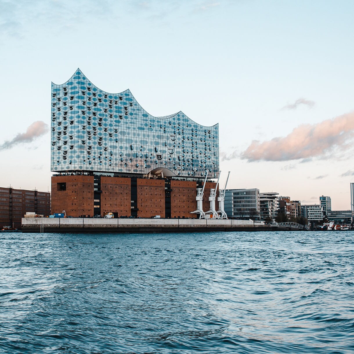 Exterior view of the Elbphilharmonie concert hall in Hamburg, featuring a glass façade shaped like waves built atop a red brick base along the waterfront.