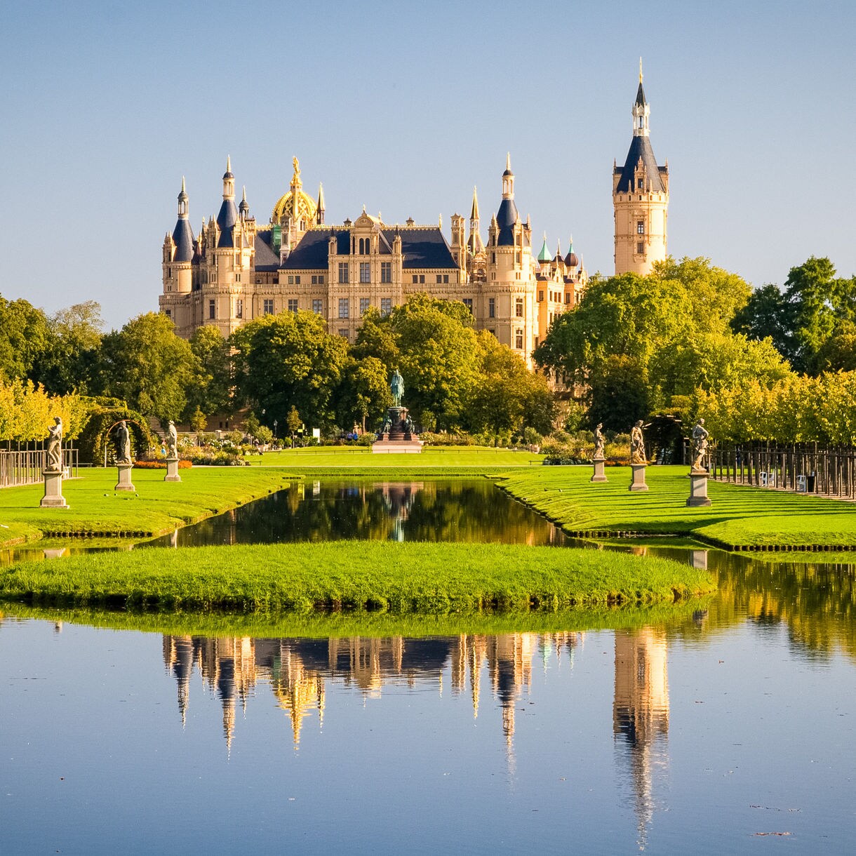 View of Schwerin Castle in Germany with ornate towers, lush gardens and a reflecting pond in front under a clear blue sky.