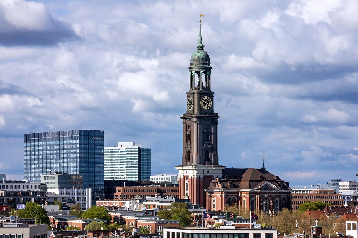 View of St. Michael’s Church in Hamburg, Germany, its tall clock tower rising against a backdrop of modern glass buildings and cloudy sky.
