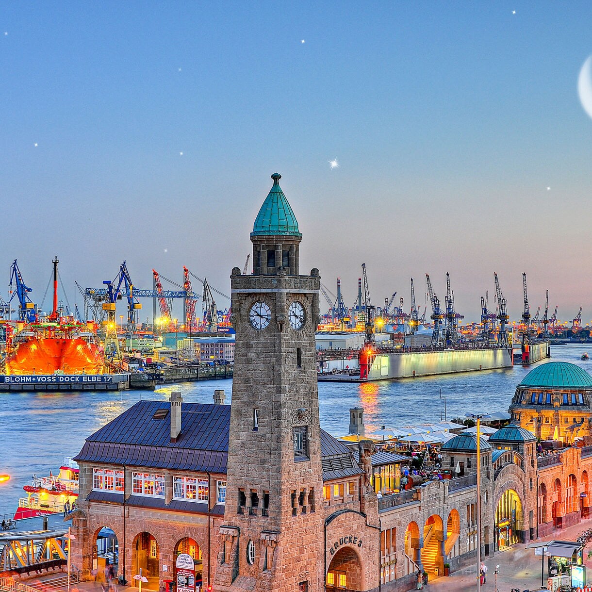 Evening view of Hamburg’s Landungsbrücken with its stone clock tower, illuminated piers and shipyard cranes glowing across the harbor under a crescent moon.