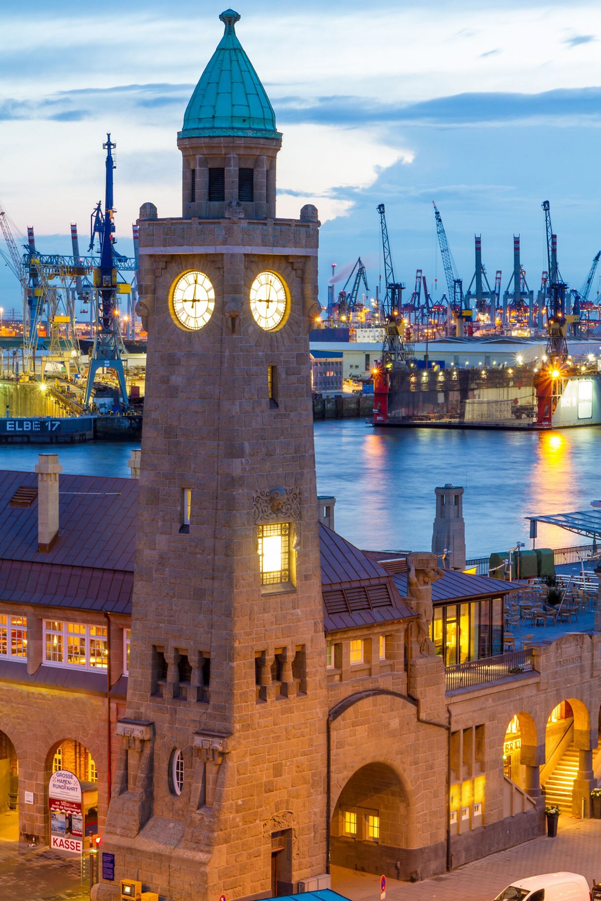 Illuminated stone clock tower at Hamburg’s Landungsbrücken with cranes, docks and ships lit up in the harbor behind it.