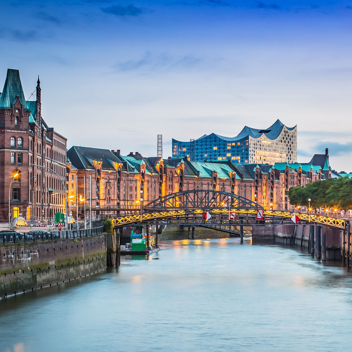 A calm canal at twilight flows beneath an arched steel bridge, lined with ornate red-brick warehouses lit in warm amber. The wavy glass Elbphilharmonie rises in the distance under a deep blue sky, with green trees along the right bank.