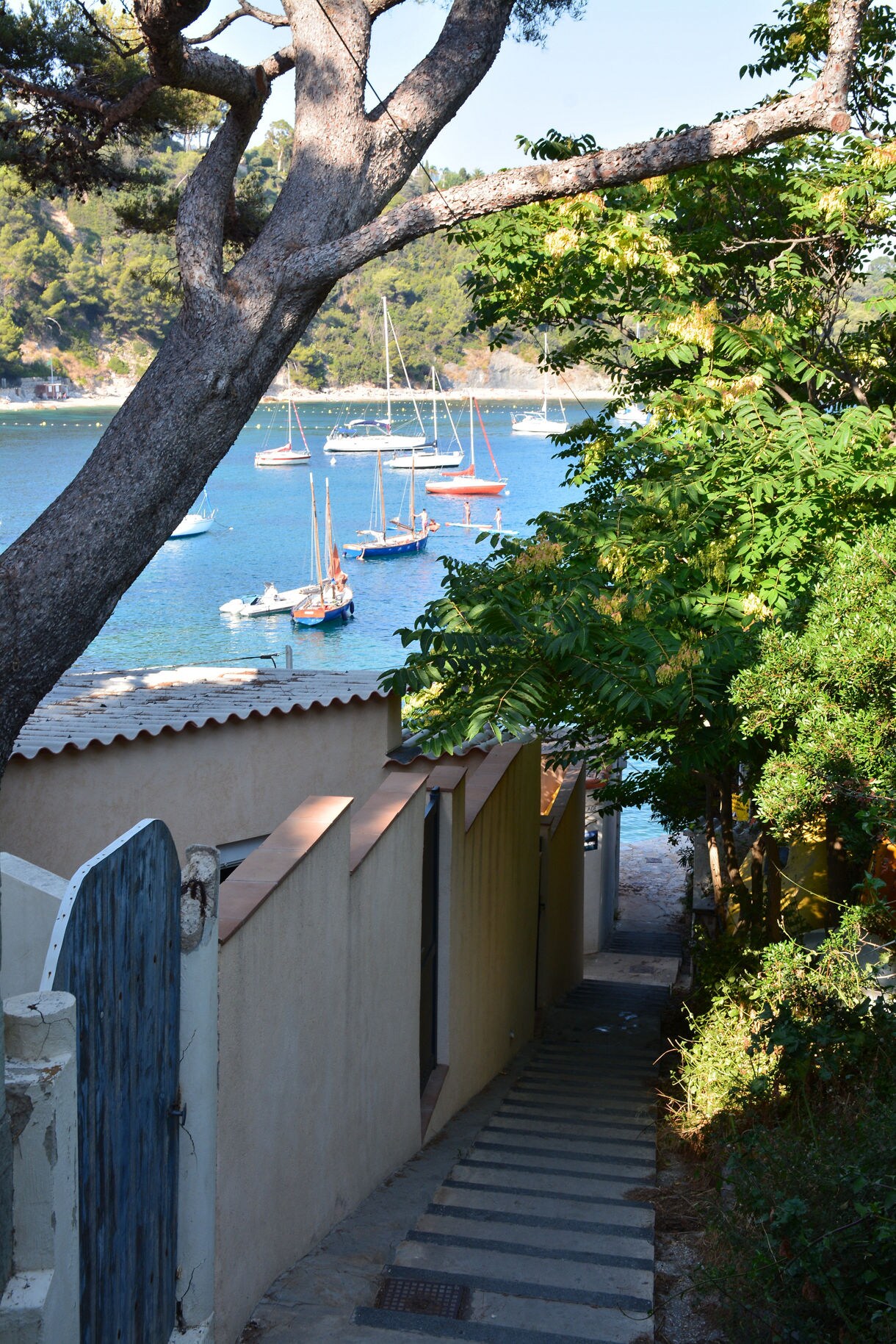 Narrow stone steps lead between stucco walls toward a small beach lined with moored sailboats on calm turquoise water, framed by trees and a large overhanging branch