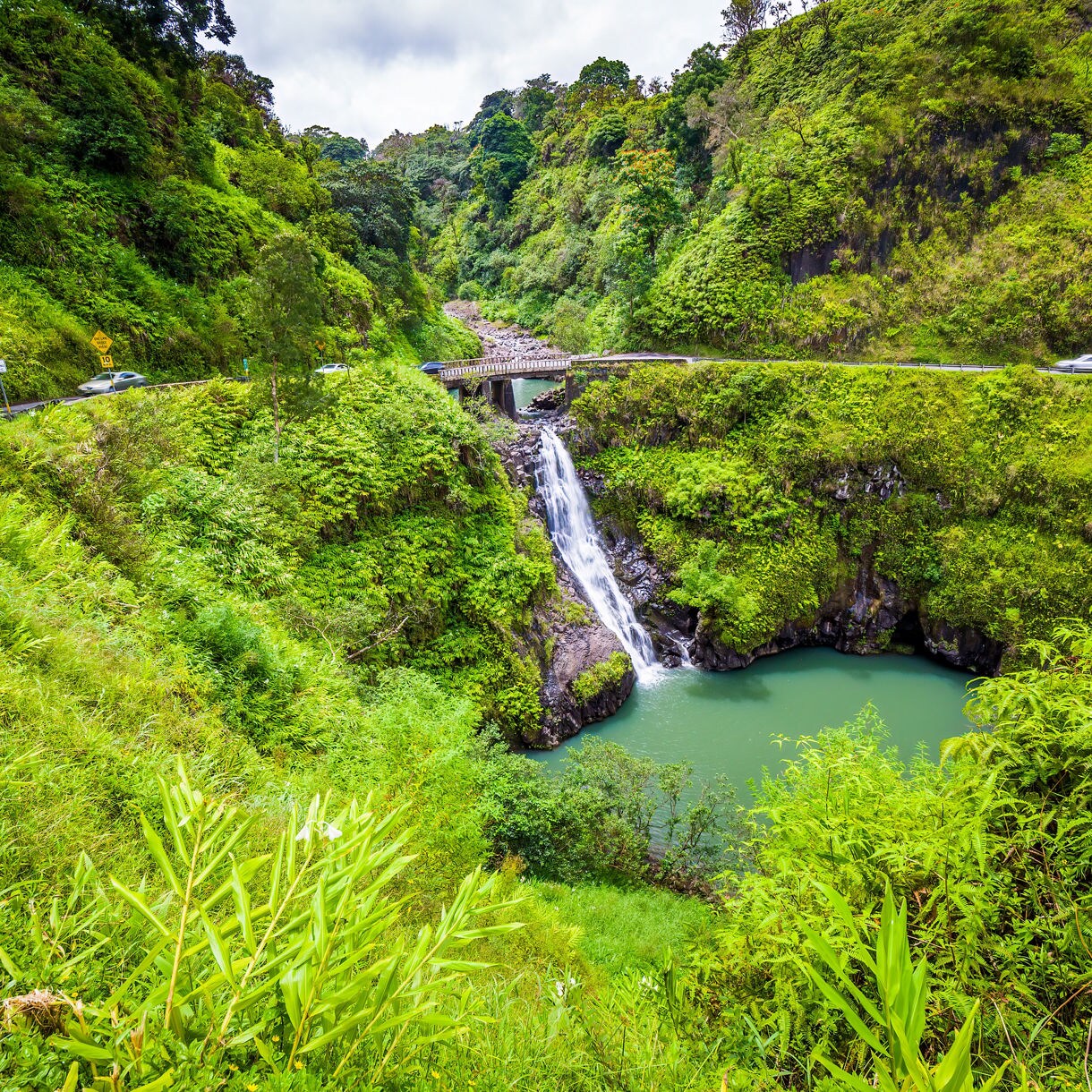 A small waterfall cascading beneath a bridge surrounded by lush green cliffs and winding roads on Maui’s Road to Hana.