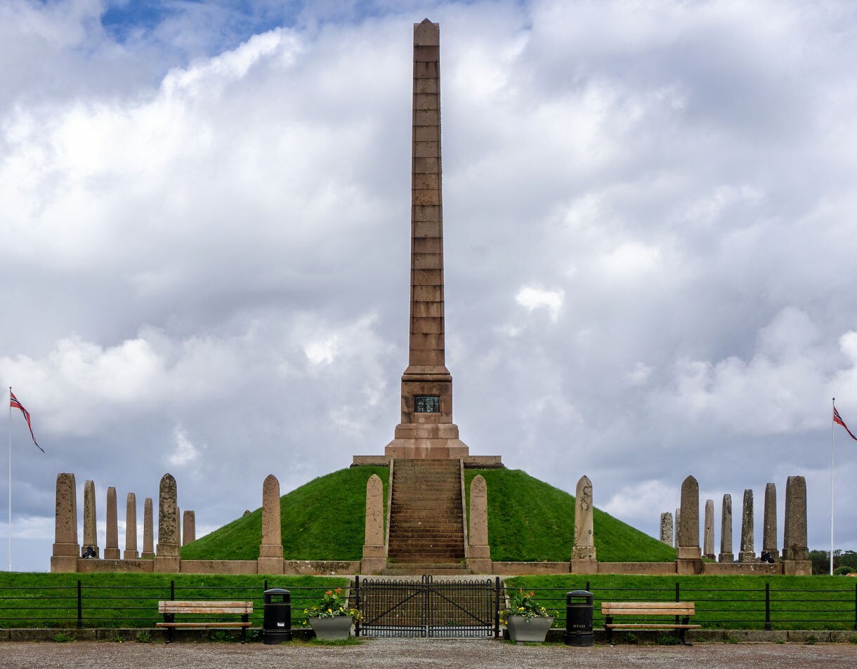 Haraldshaugen Monument in Haugesund, Norway, featuring a tall central obelisk on a grassy mound, surrounded by smaller stone pillars and Norwegian flags.