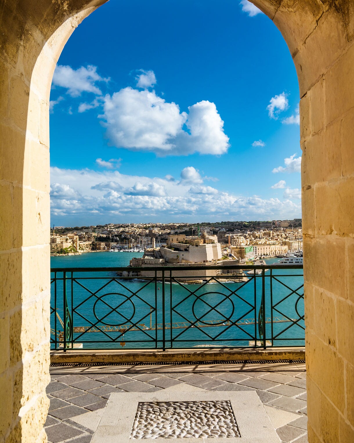 Scenic view of Malta’s Grand Harbour framed by a stone arch in Valletta, with turquoise water, historic fortifications and city buildings under a bright blue sky.