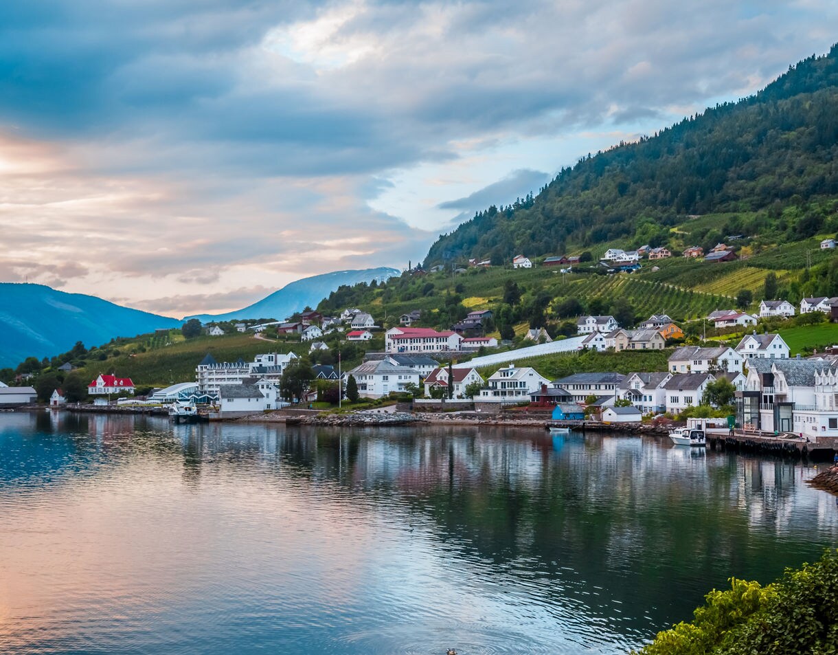 A scenic view of Lofthus village featuring white wooden houses and hillside fruit orchards along the shoreline of a tranquil fjord beneath a soft, cloudy twilight sky.