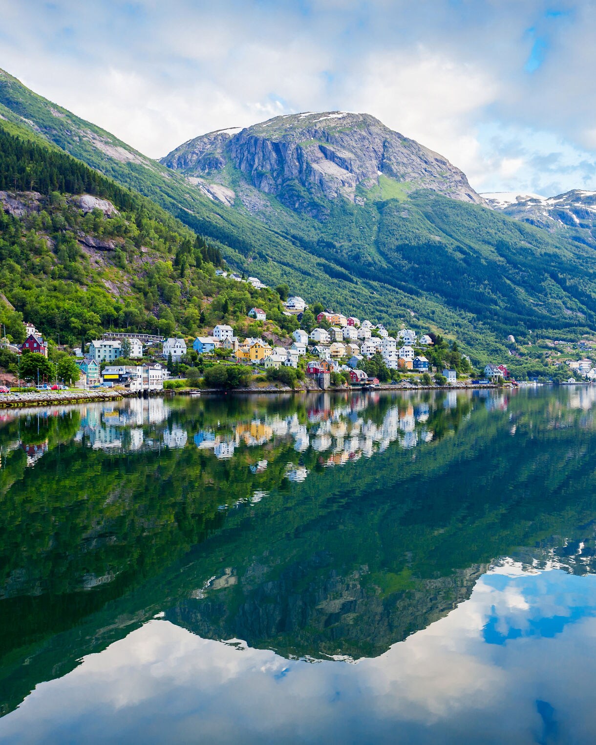 Colorful houses clustered along a lush green hillside reflect seamlessly on the calm water of a fjord beneath soaring mountains and scattered clouds.