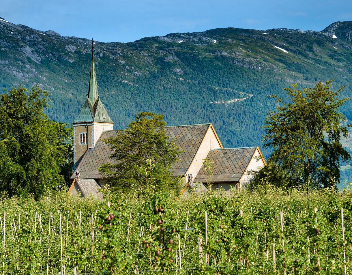 A traditional stone church with a pointed green spire rises above a dense, green fruit orchard with rocky, forest-covered mountains in the background under a clear blue sky.