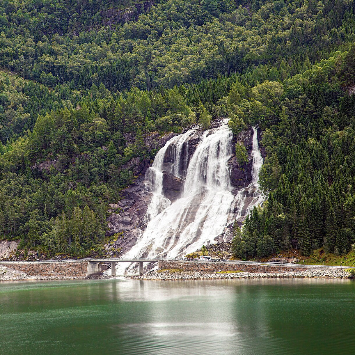 A wide waterfall cascades over rocky, tree-covered mountain slopes directly beneath a road bridge into the calm green water of a fjord.