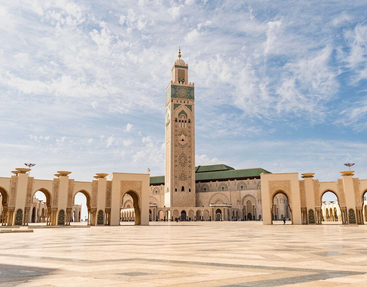 Wide plaza with elegant arches leading to a tall mosque minaret under a partly cloudy sky.