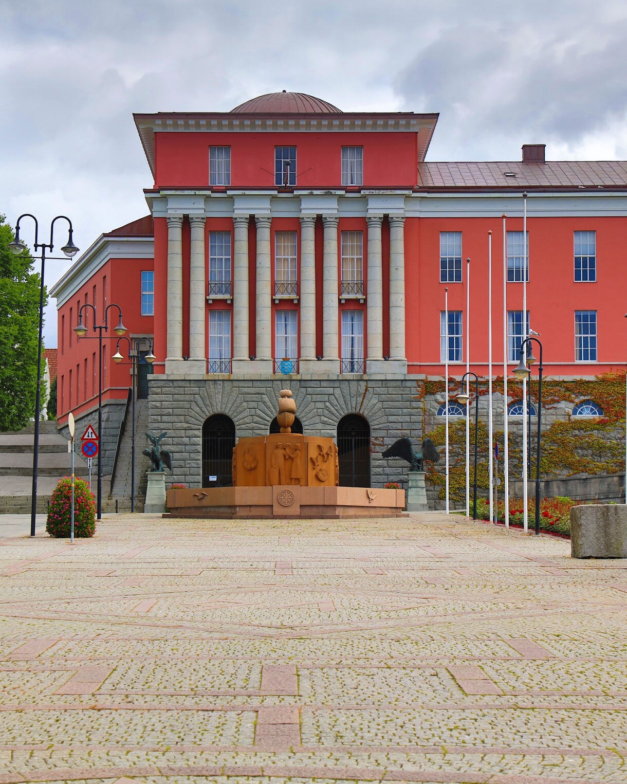 Haugesund City Hall in Norway, a bold red building with tall columns and a central fountain, framed by trees and lampposts on a cobblestone square.