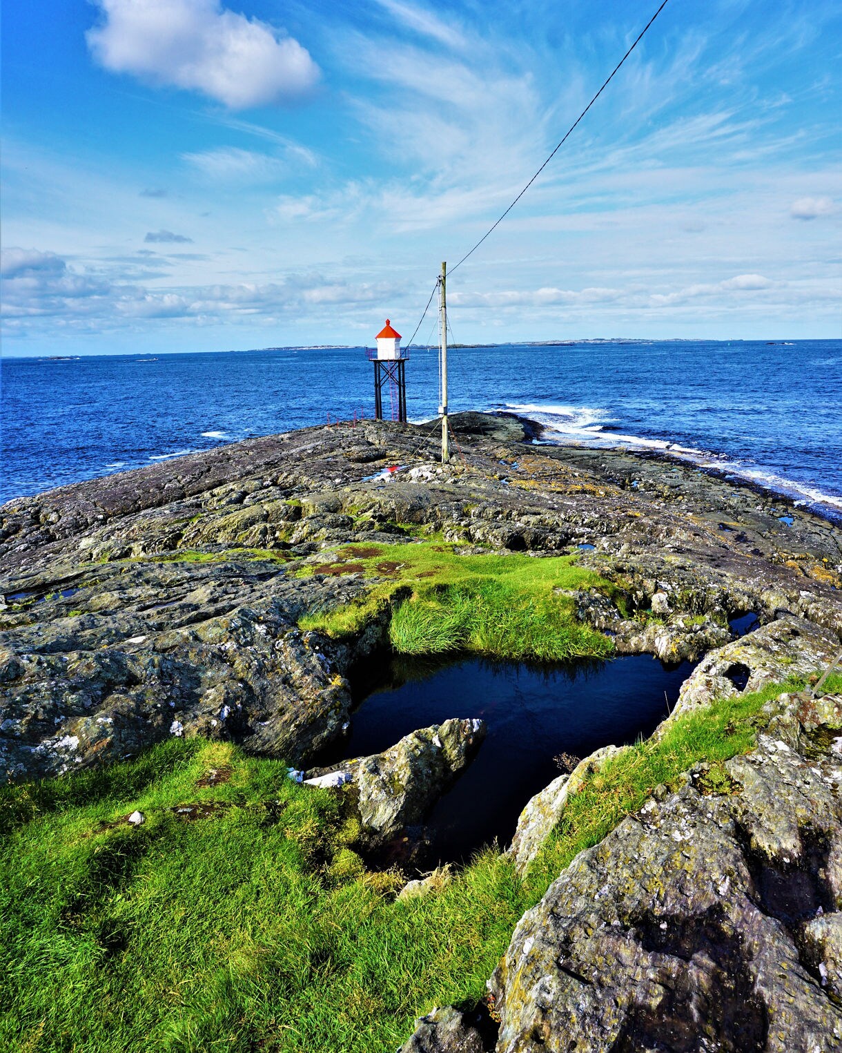 A small lighthouse with a red roof stands on rocky cliffs by the ocean in Haugesund, Norway, with patches of grass and tide pools in the foreground under a bright blue sky.