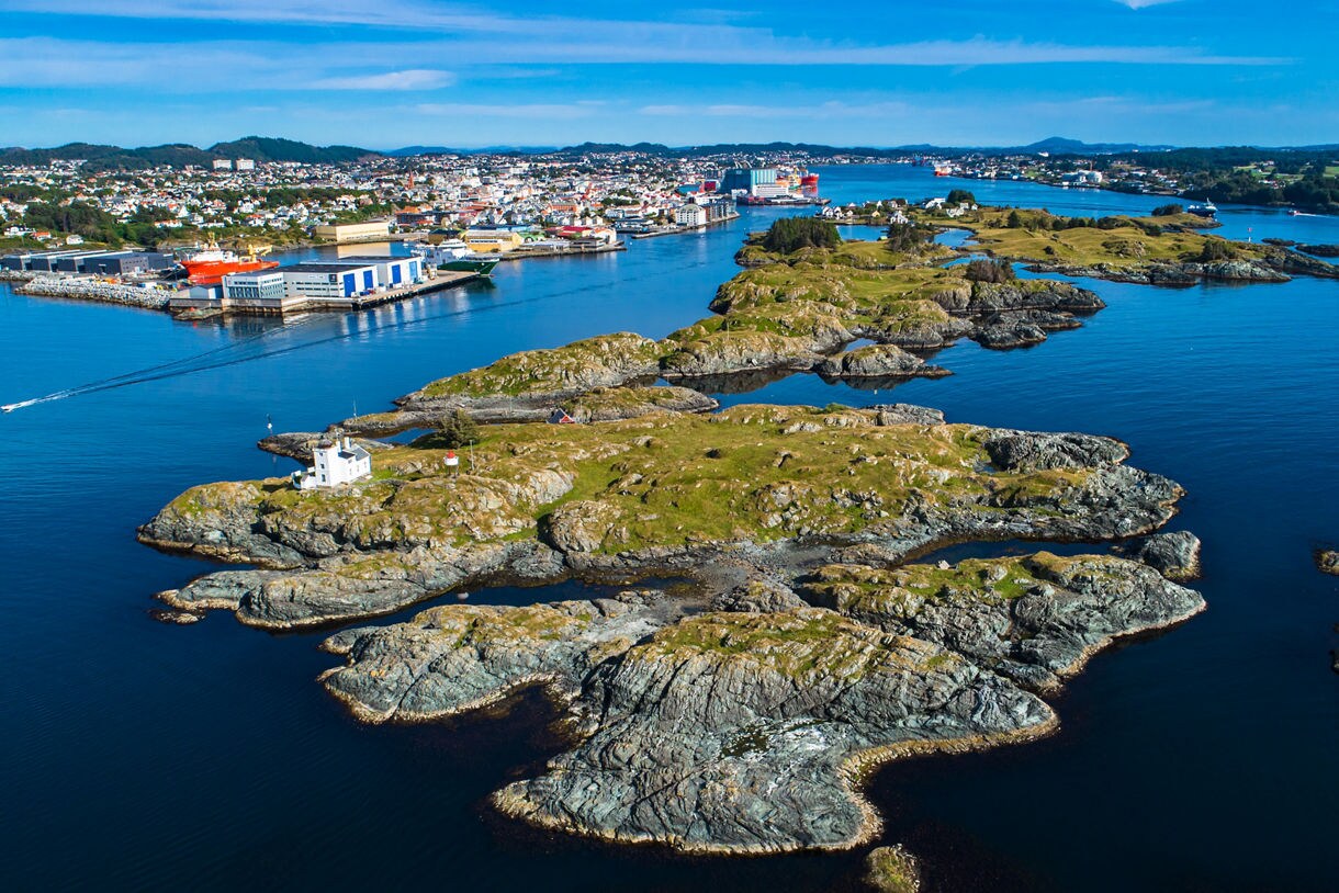 Aerial view of Haugesund, Norway, showing rocky islands in the foreground with a small lighthouse and the town’s harbor, colorful buildings and ships in the background.