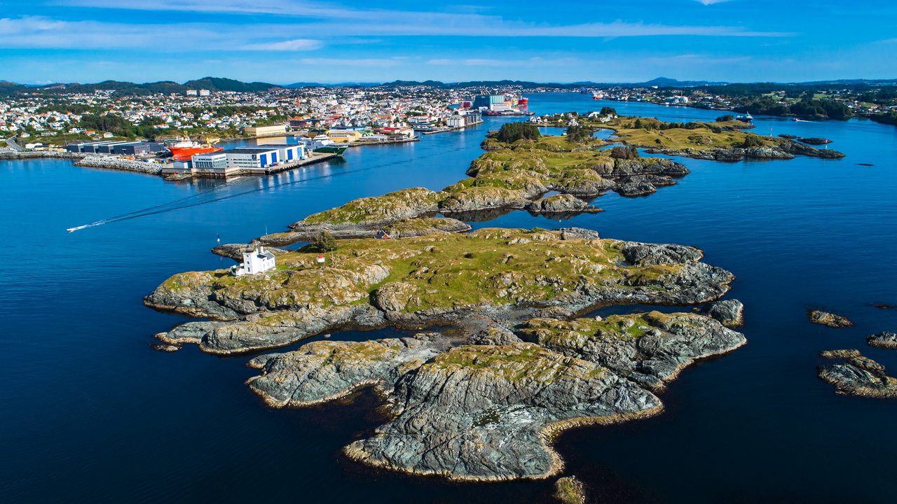 Aerial view of Haugesund, Norway, showing rocky islands in the foreground with a small lighthouse and the town’s harbor, colorful buildings and ships in the background.