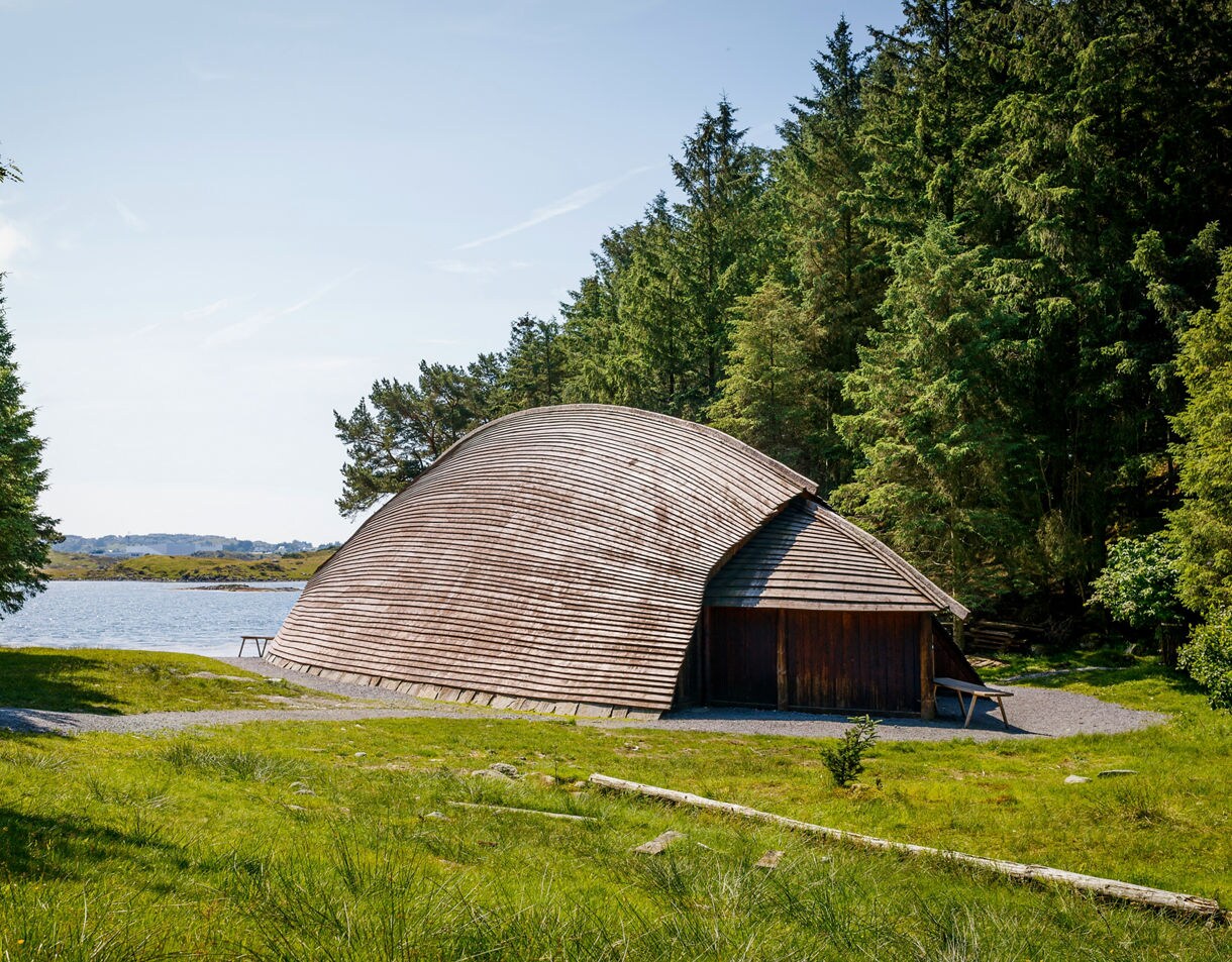 A reconstructed Viking longhouse with curved wooden roof surrounded by forest near the water in Haugesund, Norway.