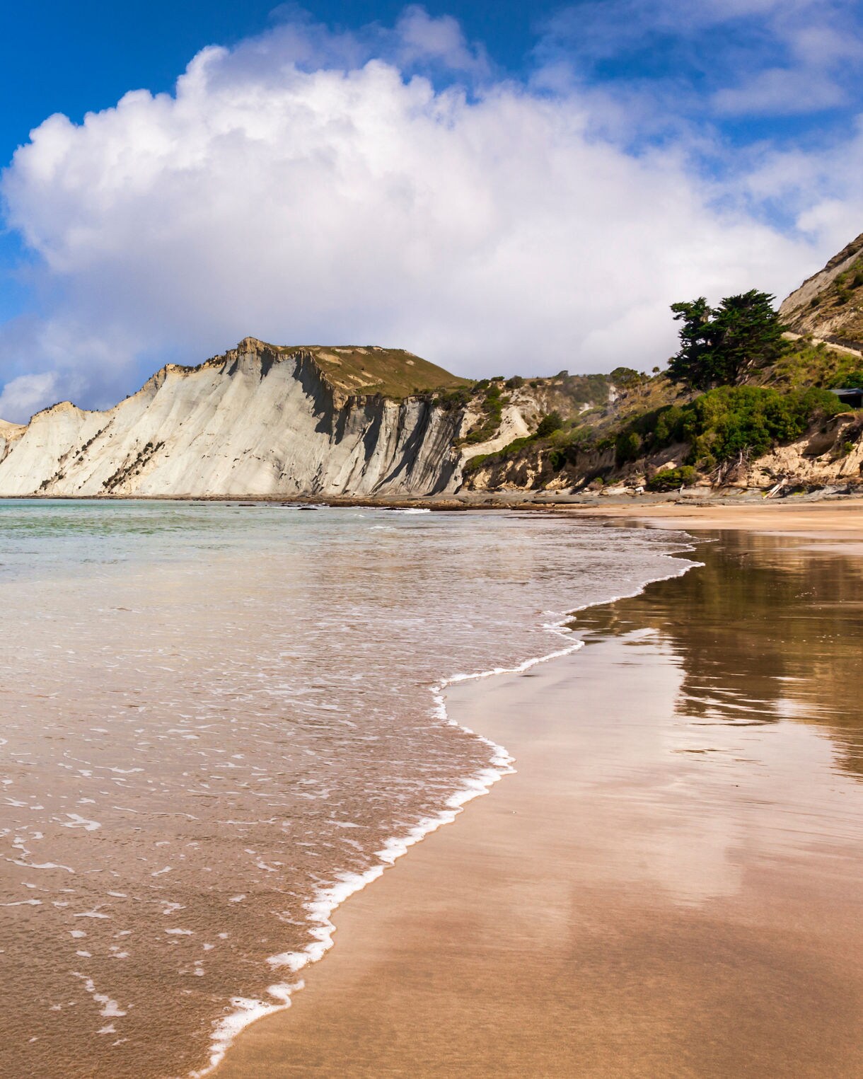 A wide beach with gentle waves lapping onto smooth golden sand, backed by tall white coastal cliffs under a bright blue sky with scattered clouds.