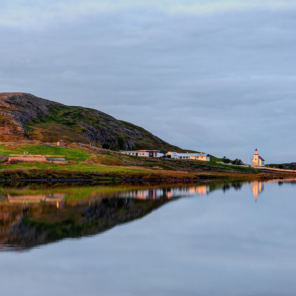 A gently sloping rocky hill and a small cluster of buildings, including a church, reflected clearly in a calm lake under soft, overcast evening light.