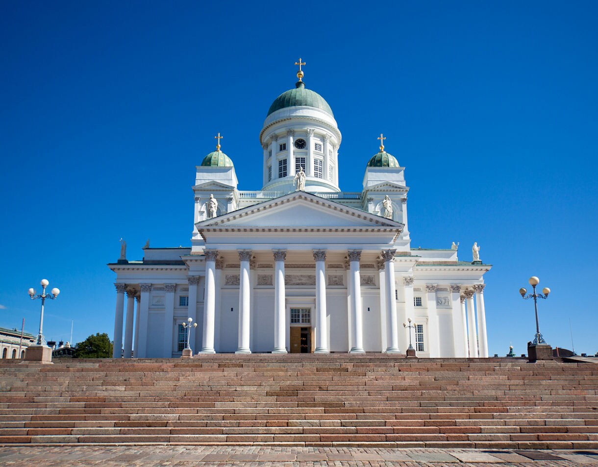 Front view of Helsinki Cathedral with tall white columns, green domes and wide stone steps leading up to the entrance under a clear blue sky.