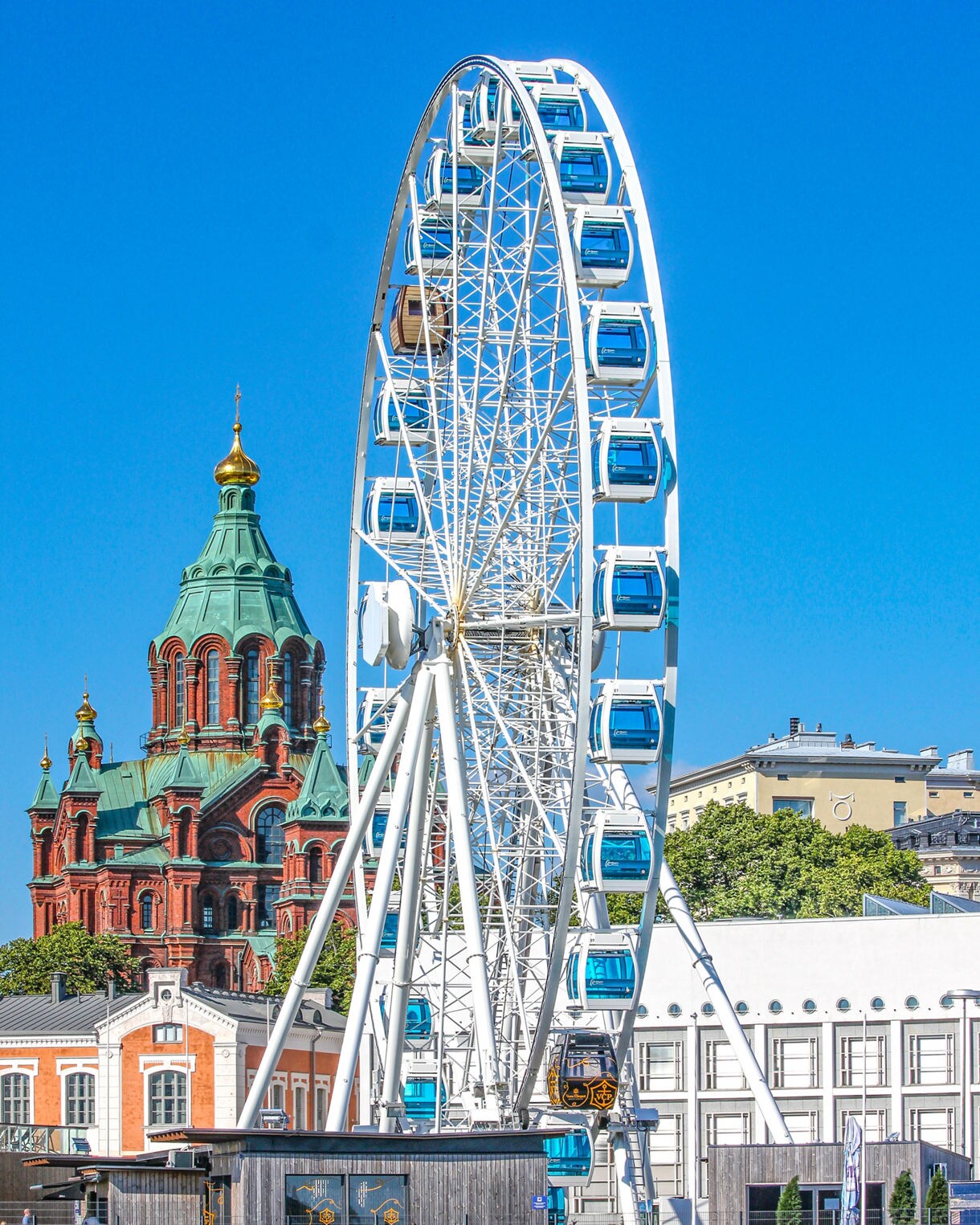 The Helsinki SkyWheel with blue gondolas standing tall by the waterfront, with the red-brick Uspenski Cathedral visible in the background under a clear blue sky.