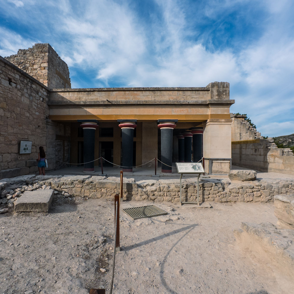 Restored section of the Palace of Knossos featuring black and red Minoan columns, stone walls and informational signs with visitors exploring the archaeological site.
