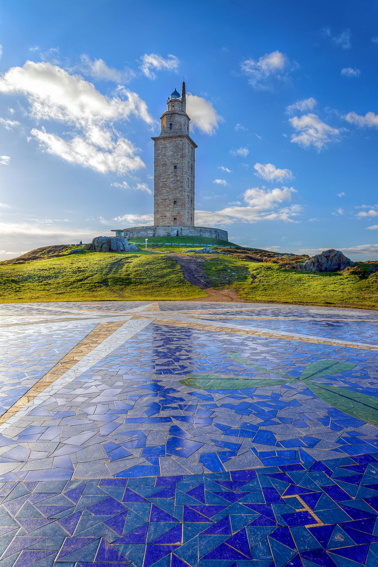 View of the Tower of Hercules on a grassy hill with a large blue compass mosaic in the foreground under a bright sky.