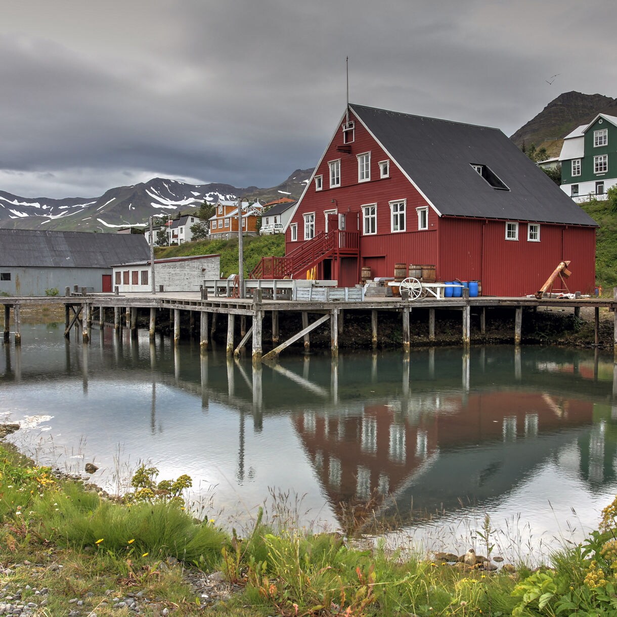 Red wooden building of the Herring Era Museum in Siglufjörður, Iceland, reflected in calm harbor water with surrounding colorful houses and snow-dusted mountains in the background.