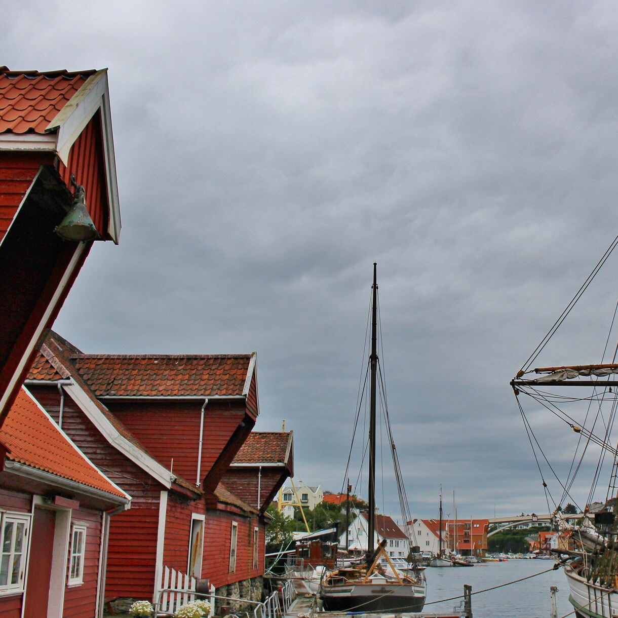 Historic red wooden buildings line the harbor in Haugesund, Norway, with sailing ships docked nearby under a cloudy sky.