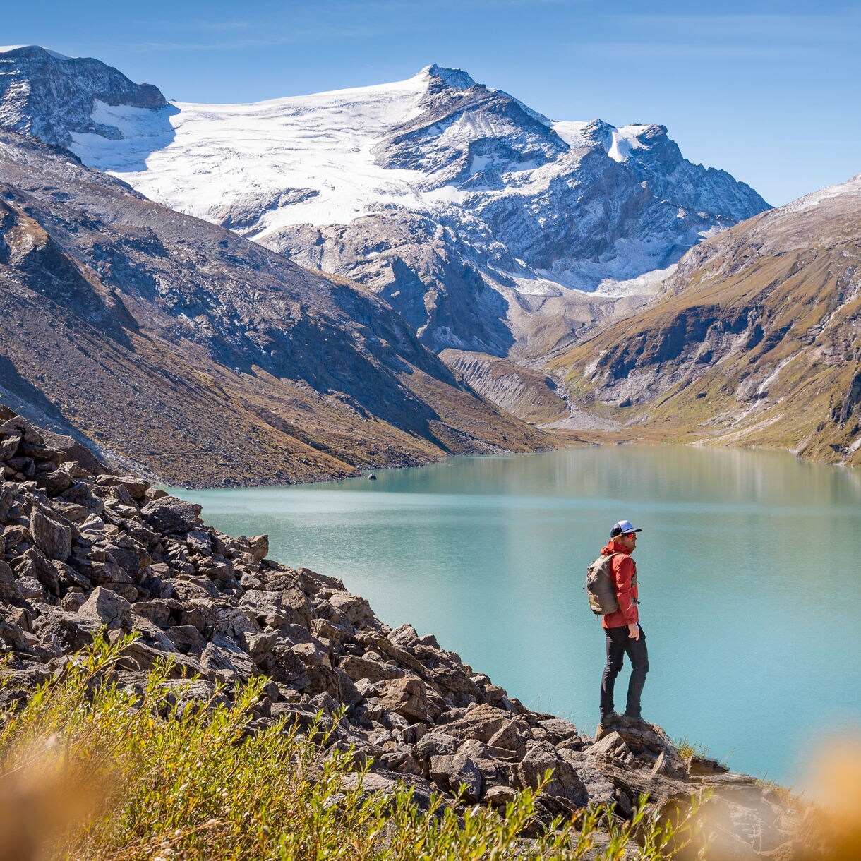 A lone hiker with a backpack stands on a rocky ridge overlooking a vivid turquoise glacial lake, surrounded by steep mountain valleys and snow-capped peaks under a clear blue sky.