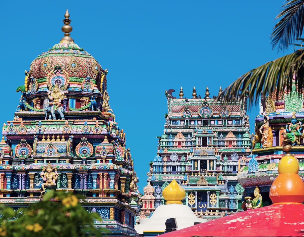 Colorful Hindu temple towers decorated with detailed carvings and statues of deities, set against a bright blue sky and framed by palm leaves.