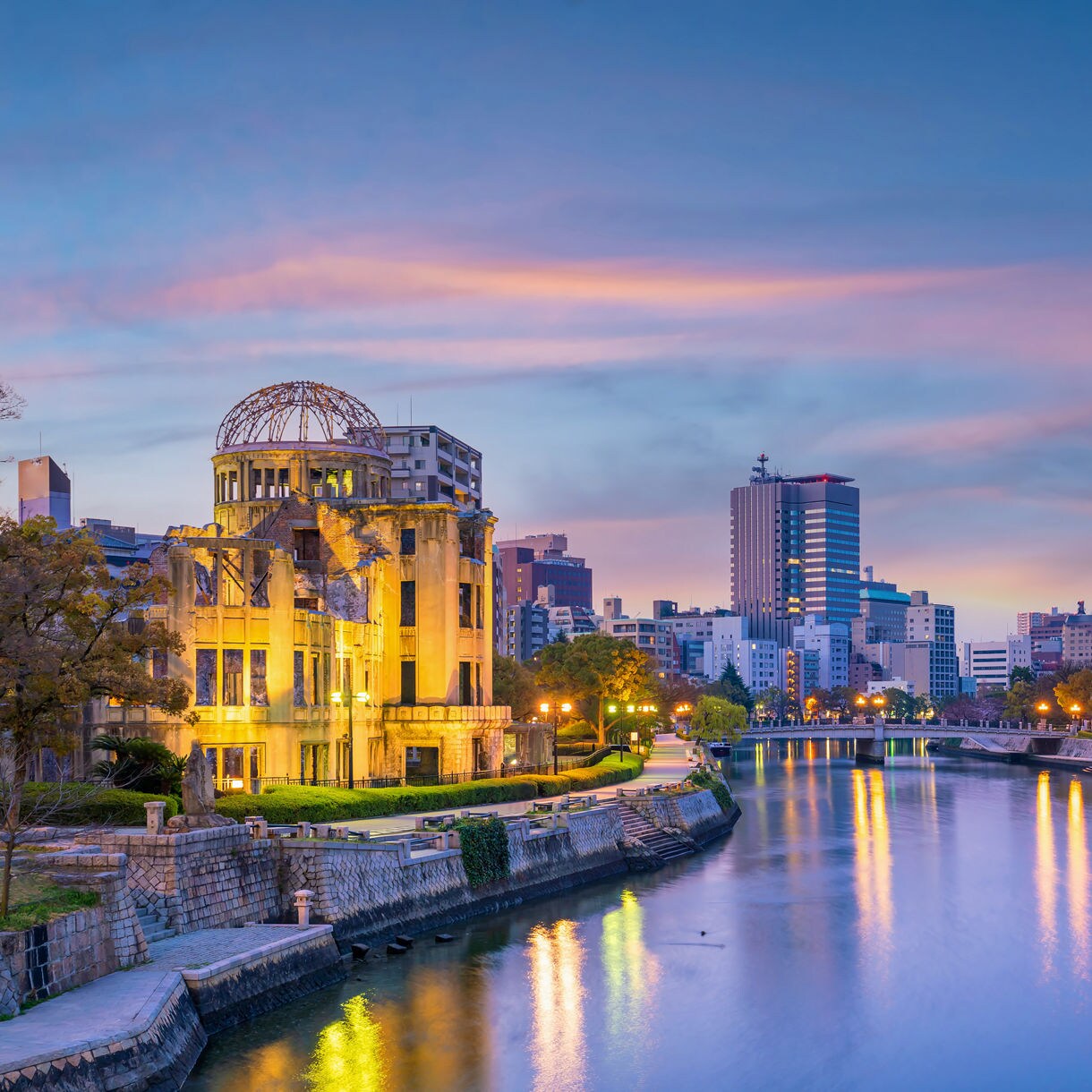 The illuminated Hiroshima Peace Memorial dome glowing beside a river reflecting city lights at dusk with the skyline in the background.