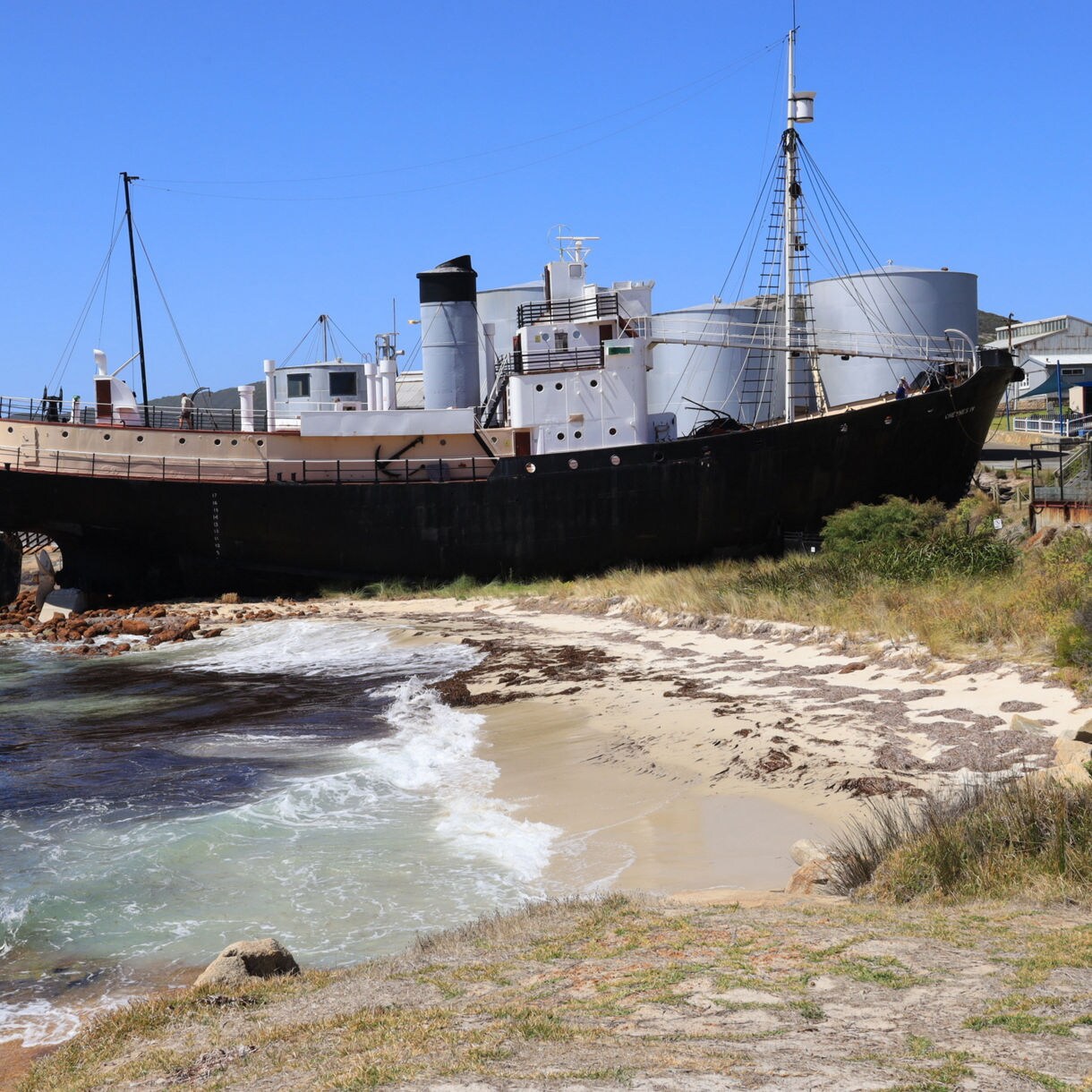 Beached black-and-tan industrial ship beside a small sandy cove with gentle waves.