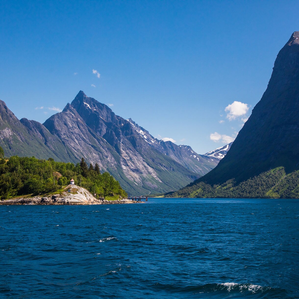 Majestic view of Hjørundfjord with sharp mountain peaks, lush green slopes and clear blue water under a bright sky in Norway.