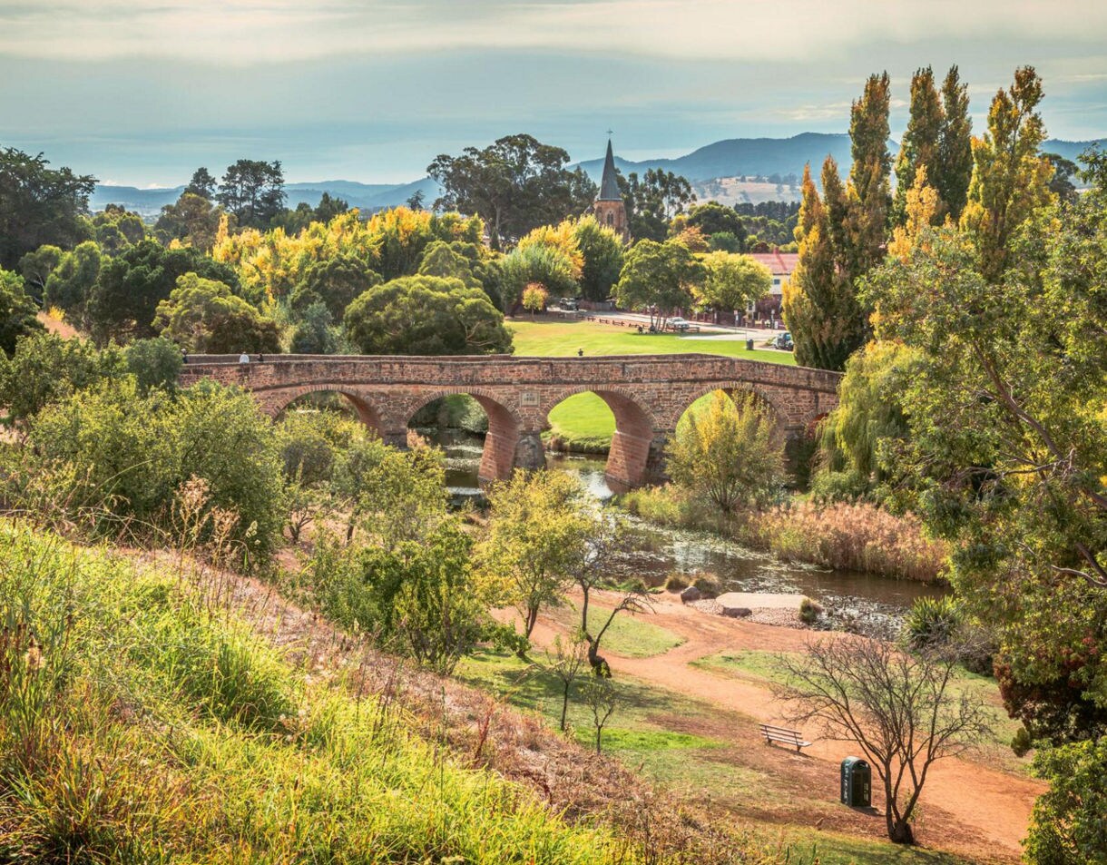 Richmond Bridge in Tasmania with stone arches spanning a river, surrounded by trees, grassy banks and a church steeple in the background.