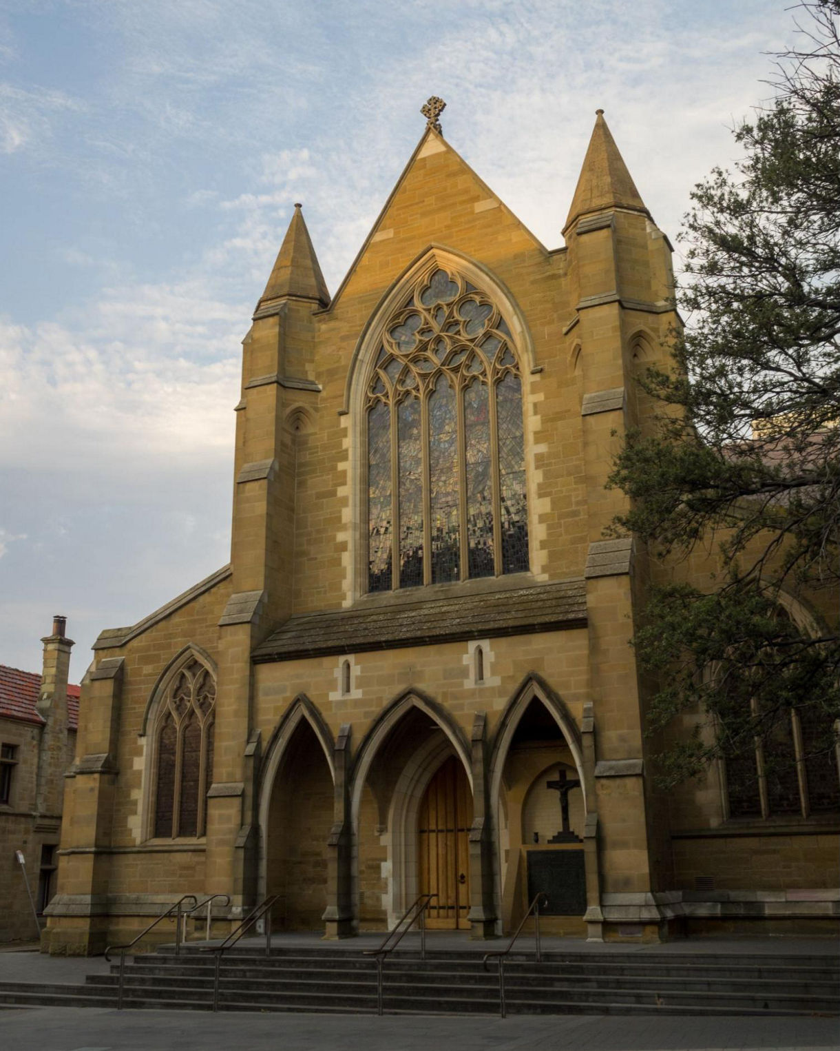Sandstone St. David’s Cathedral in Hobart with arched doorways, tall spires and a large stained-glass window under a soft evening sky.