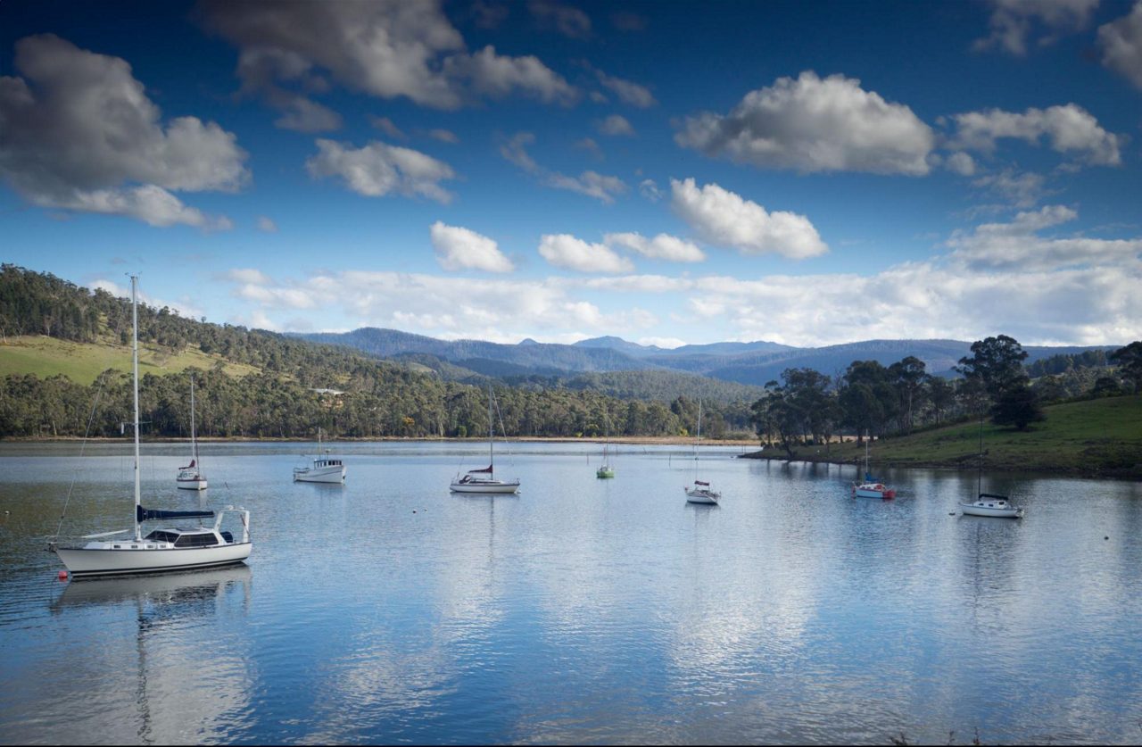 Small boats and sailboats anchored on the still Huon River in Tasmania, with forested hills and mountains in the background.