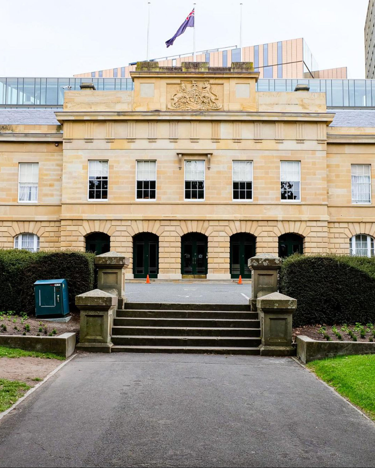 Front view of Hobart’s sandstone Parliament House, featuring neoclassical architecture with arched windows, a coat of arms crest and surrounding gardens.