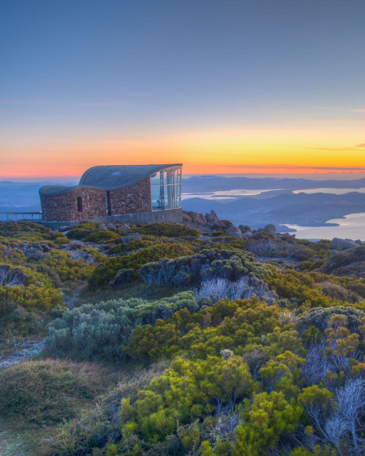 Sunrise view from Mount Wellington with a stone lookout building in the foreground and sweeping vistas of Hobart, islands and water below.