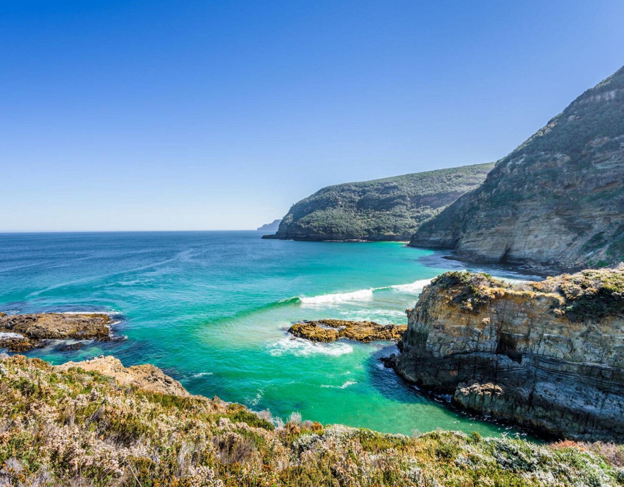 Panoramic view of the Tasman Peninsula with steep cliffs, turquoise waters, rocky outcrops and lush coastal vegetation under a clear blue sky.